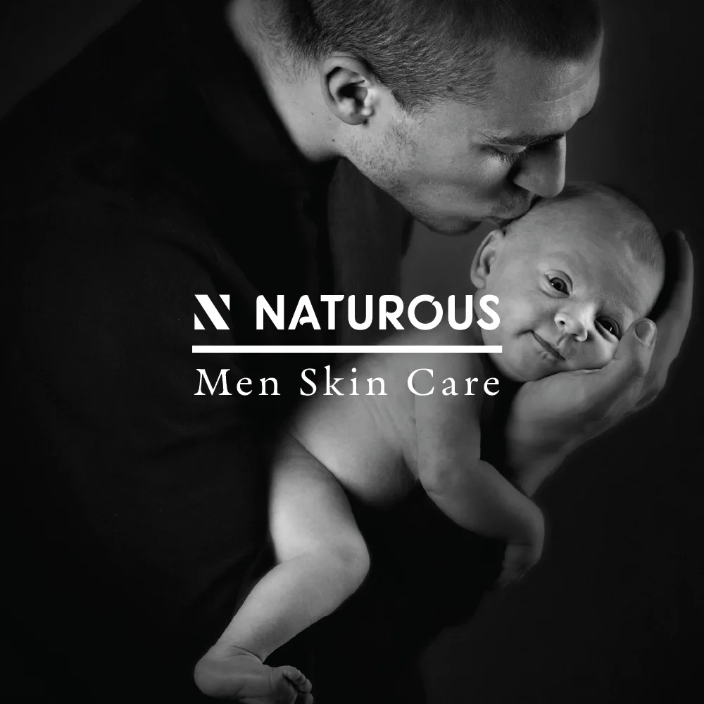 A man kisses the forehead of a baby he is holding in his hands. The image, presented in timeless black and white, bears the Naturous skincare logo centered over it with the text "NATUROUS Men Skin Care.