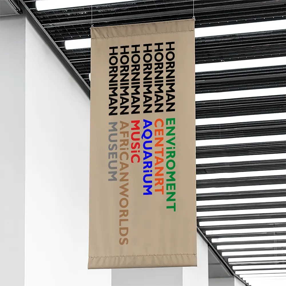 A beige vertical banner hangs from the ceiling in a modern building. The banner features "Horniman Museum" repeated vertically seven times in black text. Different topics are listed after each word in various colors: "Centarum," "Aquarium," "Music," "Africa," "Worlds," "Environment," and "Museum.