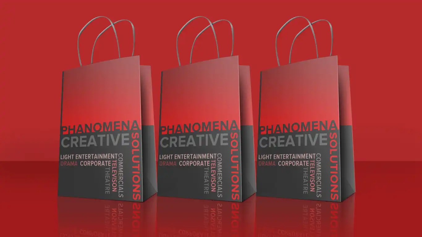 Phanomena-Exhibition-Merchandise-Design-Bags Three red and black shopping bags are displayed in a row against a red background. The text on the bags reads "PHANOMENA CREATIVE SOLUTIONS" and includes words such as "LIGHT ENTERTAINMENT," "DRAMA," "COMMERCIAL," "THEATRE," and "CORPORATE." This striking merchandise design showcases their diverse offerings.