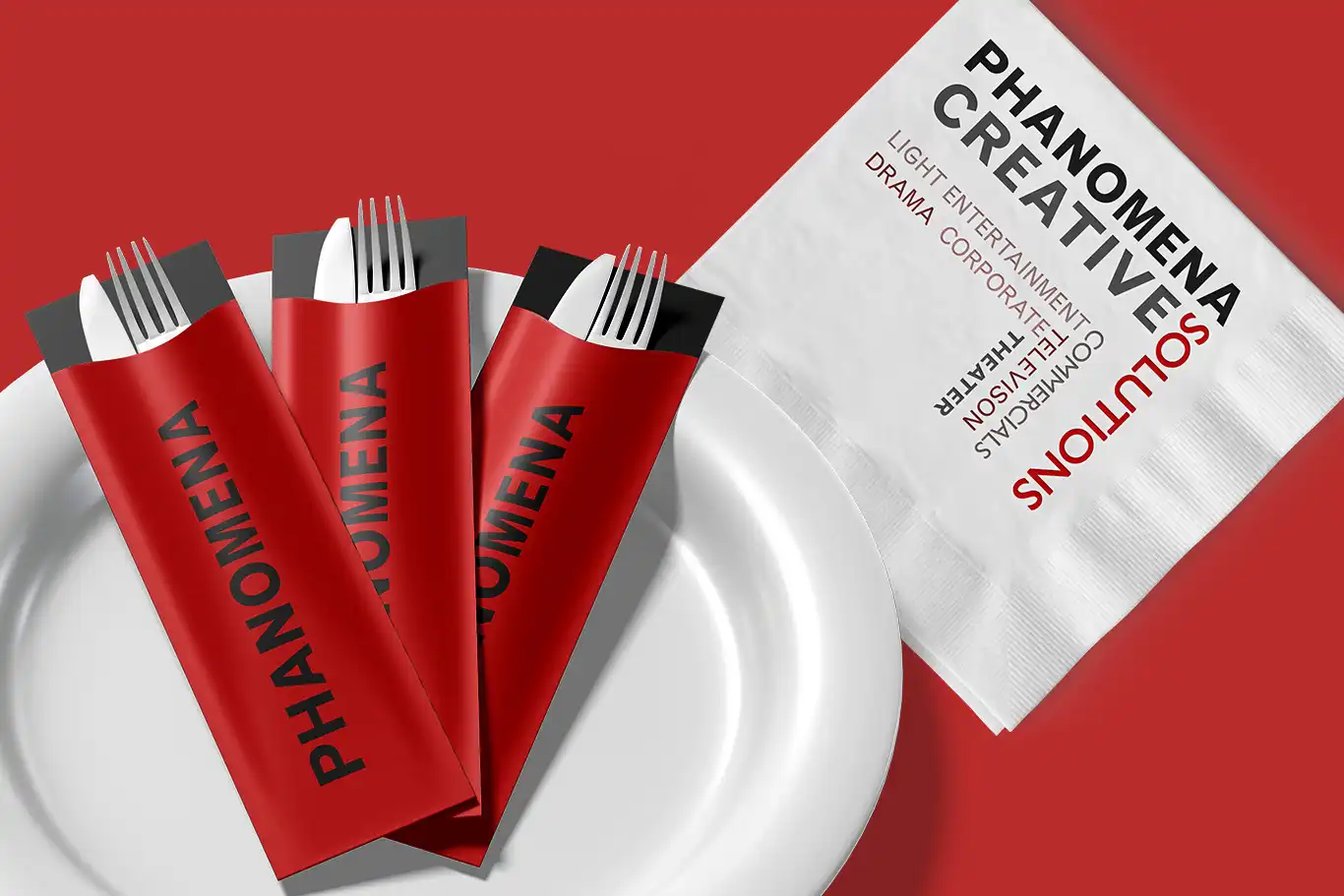 Phanomena-Exhibition-Merchandise-Design-Cutlery-and-Napkin A white plate holds three sets of cutlery wrapped in red pouches labeled "PHANOMENA." Beside the plate, a white napkin lists services such as "LIGHT ENTERTAINMENT," "DRAMA CORPORATION," "DIGITAL STRATEGIES," and more, showcasing their exceptional merchandise design.
