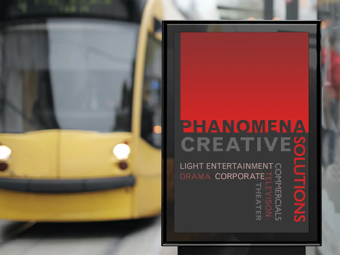 Phanomena-Exhibition-Merchandise-Design-Street-Billboard A street advertisement board displays an ad for "Phanomena Creative Solutions" offering services in light entertainment, drama, corporate, theater, commercials, television, and merchandise design. A yellow tram is visible in the blurred background.
