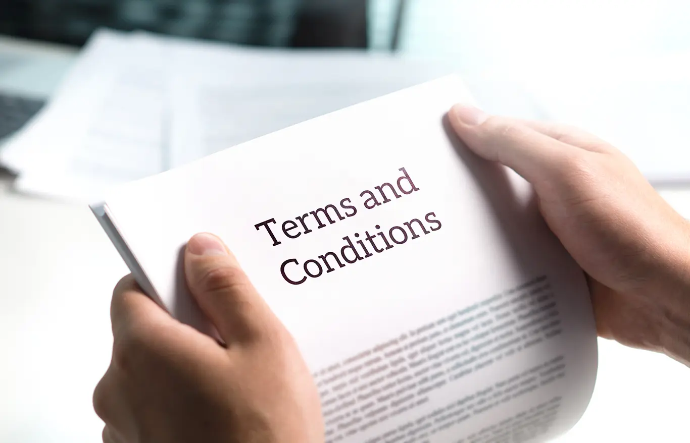 A person is holding a document titled "Terms and Conditions" printed in bold letters on the cover. The document appears to be several pages long, and blurred text is visible on both the current and underlying pages. The background is slightly out of focus. A person is holding a document titled "Terms and Conditions" printed in bold letters on the cover. The document appears to be several pages long, and blurred text is visible on both the current and underlying pages. The background is slightly out of focus.